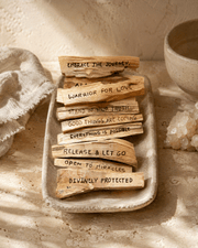 Palo Santo Mantra Sticks are natural light-brown wood pieces with rough grain, each marked with a different affirmation.