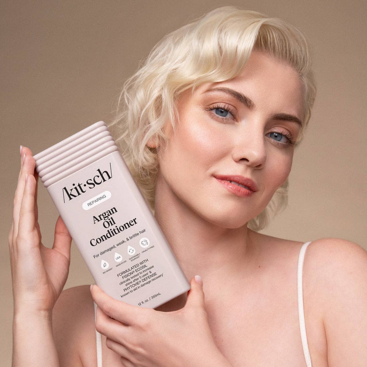 The Rooted Home Kitsch Argan Oil conditioner bottle held by a blonde woman with a soft, natural makeup look.