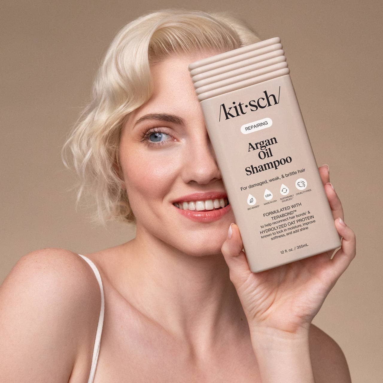 The Rooted Home Kitsch Repairing Argan Oil Shampoo bottle held by a woman with blonde hair against a neutral background