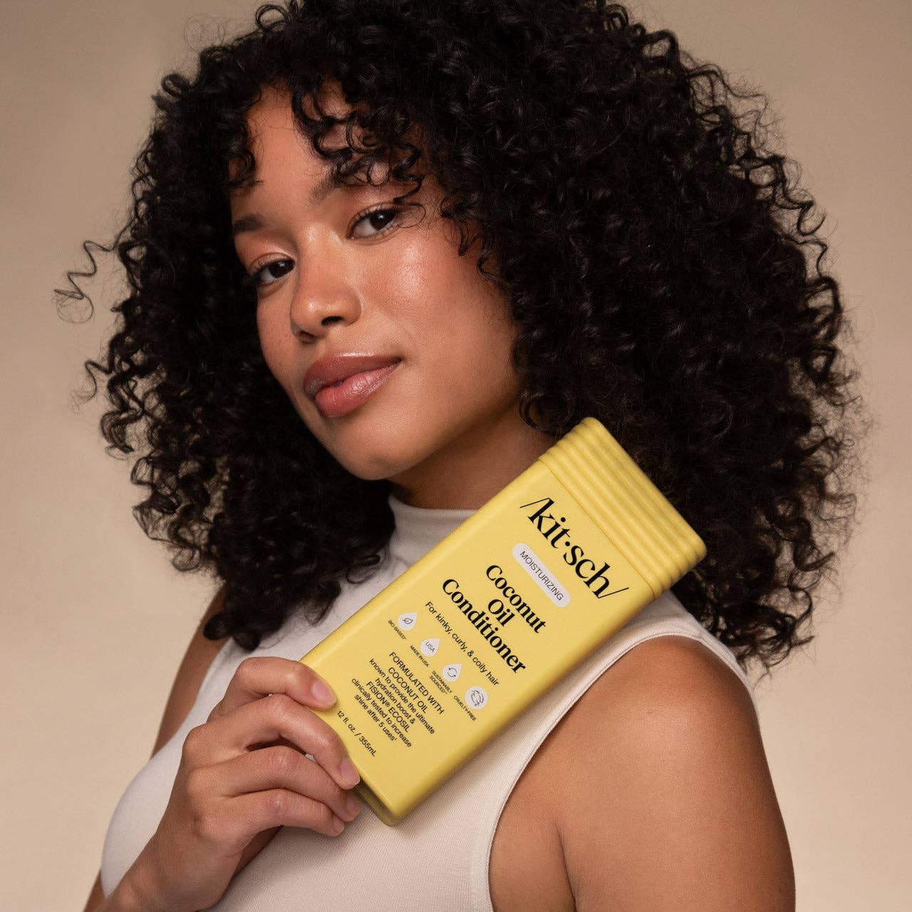 The Rooted Home Kitsch moisturizing coconut oil conditioner bottle held by a woman with curly hair