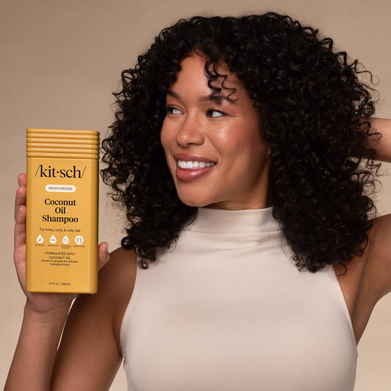 The Rooted Home Kitsch moisturizing coconut oil shampoo bottle held by a smiling woman with curly hair